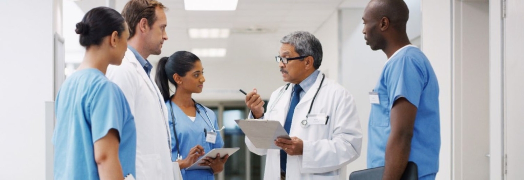 A diverse group of health-care professionals, including doctors and nurses, stand in a hospital corridor. The group is engaged in discussion, with one male doctor holding a clipboard and gesturing as others listen attentively.