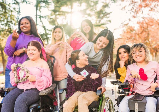 Eight girls, four in wheelchairs, chat and laugh under trees in dappled sunlight.