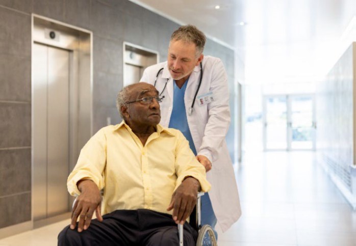 An elderly African American man is being pushed through a health-care facility in a wheelchair by a health-care professional