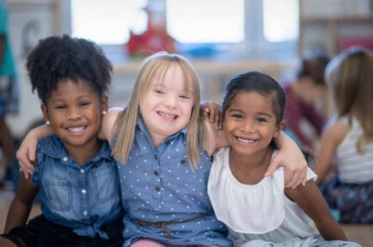 Three young girls from diverse backgrounds sit with their arms over eachother's shoulders, smiling into the camera.