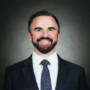 A formal photograph of Dr. Carney in a dark suit, white, shirt, and blue tie. He has dark hair and a beard.